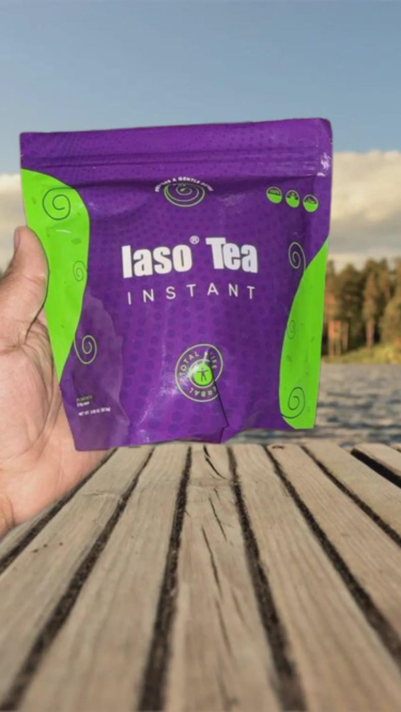 May include: A purple and green packet of "Iaso Tea Instant" held in a hand. The packet has green swirls and the words "Iaso Tea" in white. The background shows a lake and trees.
