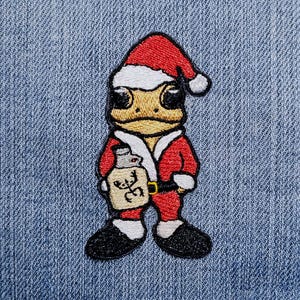 Könnte beinhalten: Gestickter Aufnäher mit einem Frosch als Weihnachtsmann, der einen roten Anzug und Hut mit weißem Besatz trägt. Der Frosch hält eine Flasche mit einem Hirschmotiv. Der Aufnäher befindet sich auf einem blauen Jeanshintergrund.