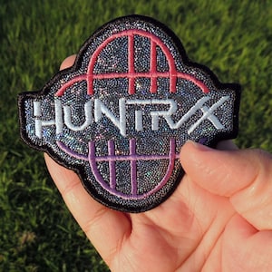 May include: A black, glittery patch with the word "HUNTRAX" in white, outlined in silver. The patch features a pink and purple design. The patch is held in a hand against a background of green grass.