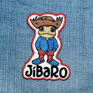 Könnte beinhalten: Gestickter Aufnäher mit einer Cartoon-Froschfigur, die einen braunen Cowboyhut, ein blaues Hemd, rote Shorts und blaue Stiefel trägt. Der Aufnäher hat einen weißen Rand mit roter Umrandung und das Wort "JíBARO" in schwarzen Buchstaben.