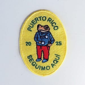 Könnte beinhalten: Gestickter gelber Aufnäher mit einem Frosch in einer blauen Jacke und roter Hose. Der Text "PUERTO RICO" und "SEGUIMO AQUI" ist grün. Die Zahlen "20" und "25" sind ebenfalls vorhanden.