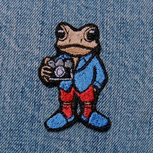 May include: Embroidered patch featuring a cartoon frog holding a camera. The frog is wearing a blue shirt, red pants, and blue shoes. The patch has a black border and is set against a denim background.
