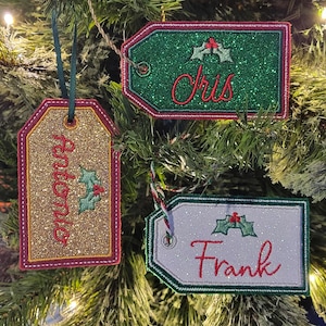 May include: Three Christmas tree ornaments in the shape of gift tags. One tag is gold glitter with the name "Antonio" embroidered in red. Another is green glitter with "Chris" in red. The third is white with "Frank" in red.