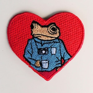 Könnte beinhalten: Gestickter Aufnäher in Form eines roten Herzens. Der Aufnäher zeigt einen Frosch mit blauem Hemd, der einen weißen Becher und eine Kamera hält. Der Frosch ist hellbraun und das Herz ist rot.