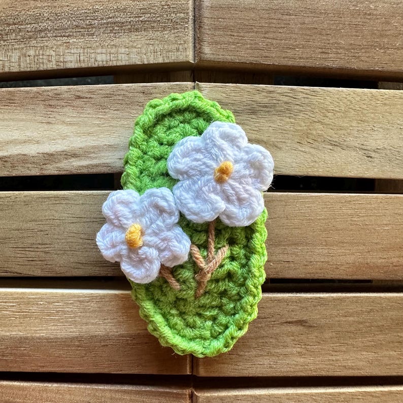 May include: A handmade crochet floral applique featuring two white flowers with yellow centers on a green leaf. The leaf and flowers are made of yarn, and the applique is set against a wooden background.