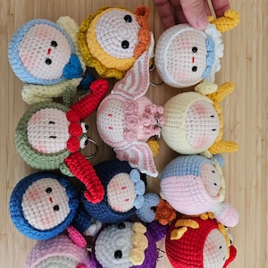 May include: A collection of colorful, handmade crochet doll keychains. Each keychain features a round head with black button eyes and a small red mouth. The dolls are in various colors, including blue, yellow, pink, and red.