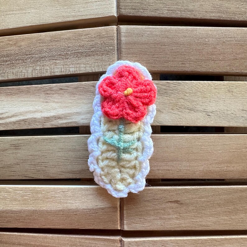 May include: A handmade crochet flower pin with a red flower and yellow center. The flower is attached to a white and cream-colored base with a green stem. The pin is set against a wooden background.