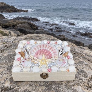 Puede incluir: Una caja rectangular de color beige con un cierre metálico de bisagra, con una tapa decorada con un surtido de conchas marinas blancas y rosas, pequeñas estrellas de mar y perlas blancas dispuestas en un patrón denso y superpuesto.