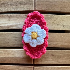 May include: A handmade, crochet hair clip featuring a vibrant pink base with a white and yellow flower accent. The flower is centered on the pink oval-shaped clip, creating a charming, decorative accessory.