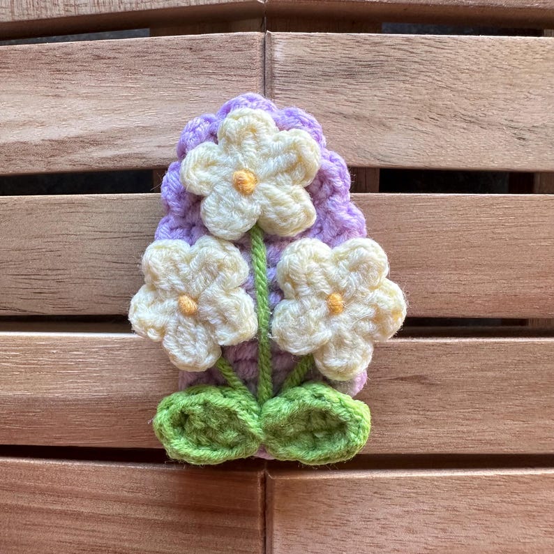 May include: A handmade crochet flower pin featuring three cream-colored flowers with yellow centers, green stems and leaves, and a lavender backing. The pin is set against a wooden background.