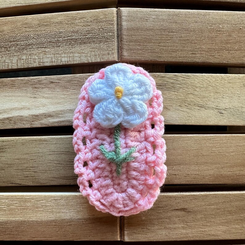 May include: A handmade crochet applique in the shape of a pink oval with a white flower and green stem. The flower has a yellow center. The applique is made of yarn and is likely used for embellishing clothing or accessories.