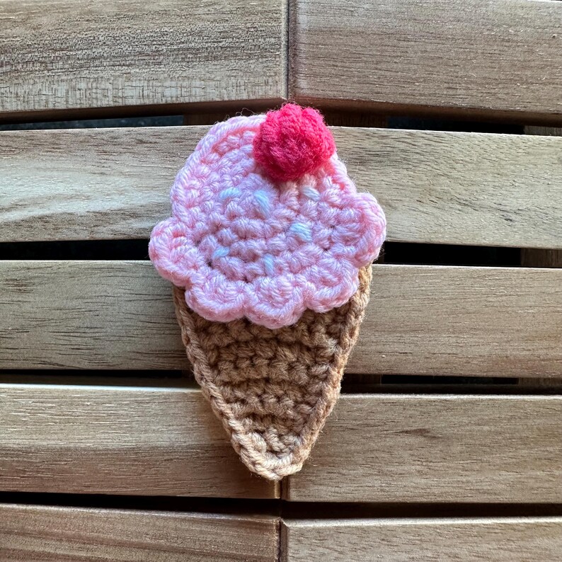 May include: A handmade crochet ice cream cone. The ice cream is pink with a white swirl and a bright pink cherry on top. The cone is brown. The craft is displayed on a wooden surface.
