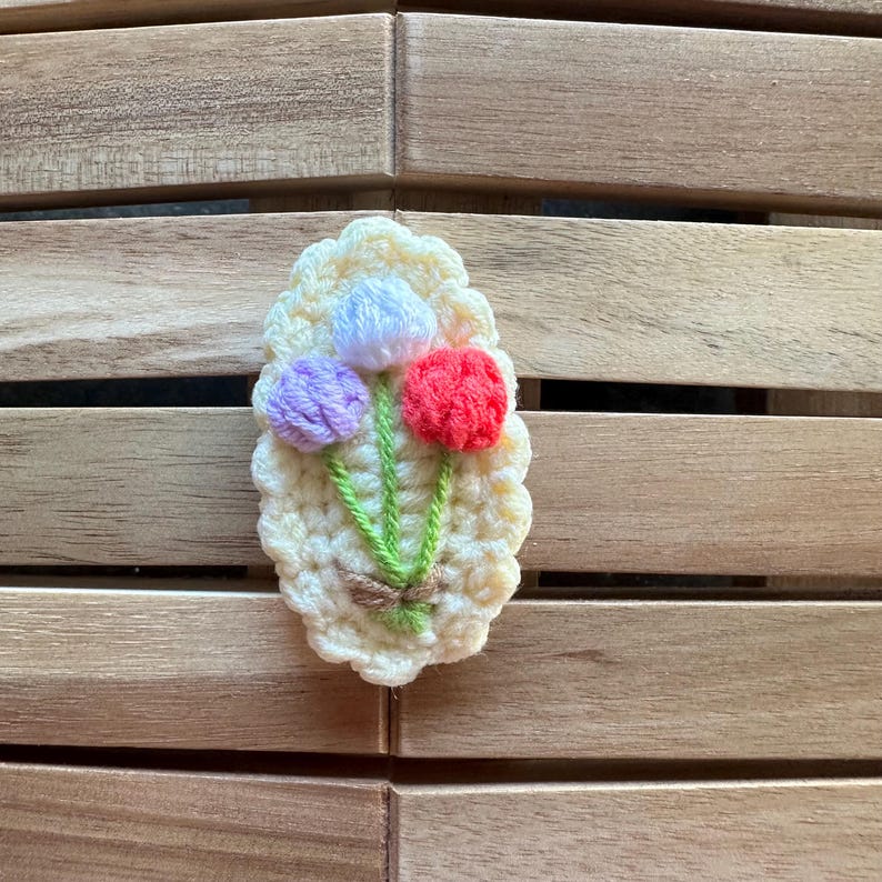 May include: A handmade crochet flower brooch featuring three colorful flowers: purple, red, and white. The flowers are attached to green stems and a beige base with a scalloped edge. The brooch is set against a wooden background.
