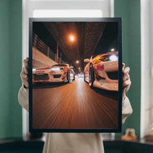 Puede incluir: Impresión enmarcada con dos coches de drift blancos y rosas corriendo de noche. Los coches tienen gráficos personalizados y se mueven hacia el espectador. La imagen está ambientada en un puente y luces de la ciudad.