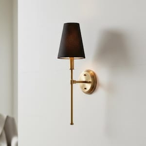 Puede incluir: Aplique de pared con pantalla negra y acabado en latón. El aplique tiene un brazo largo y delgado que se extiende desde una base circular. La pantalla tiene forma de cono y dirige la luz hacia abajo.