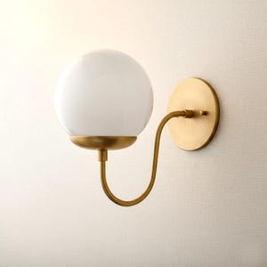 May include: A wall-mounted light fixture featuring a white globe shade and a curved, brass-colored arm. The fixture has a circular base and a brass finish, creating a modern and elegant aesthetic. The light is designed for indoor use.
