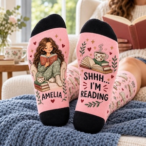Op de afbeelding: Een paar roze sokken met zwarte tenen en hielen. De linkersok toont een illustratie van een lezend persoon met een stapel boeken en de naam AMELIA. De rechtersok toont een stapel boeken, een koffiekopje en de tekst SHHH... I'M READING.