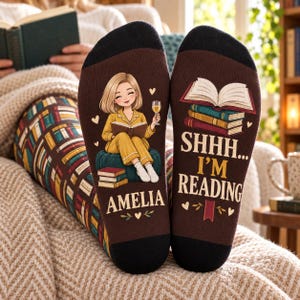 May include: A pair of brown socks with black cuffs and heels. The left sock features a cartoon of a person reading a book while holding a glass of wine, with the name AMELIA printed below. The right sock shows a stack of books and the text SHHH... I'M READING.