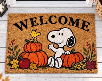 May include: A rectangular tan coir doormat featuring the black text WELCOME at the top. The design includes a white Snoopy character sitting next to orange pumpkins, a yellow Woodstock bird, a red apple, autumn leaves, and a corn cob.