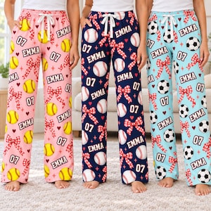 May include: Three pairs of lounge pants with white drawstring waistbands. The left pair is pink with yellow softballs, red bows, hearts, and the text EMMA and 07. The center pair is navy with baseballs, red bows, and the text EMMA and 07. The right pair is light blue with soccer balls, red bows, and the text EMMA and 07.