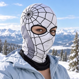 May include: A white balaclava featuring a black spiderweb pattern across the head and face, with a black-trimmed eye opening. The fabric includes a solid black neck section and a grey side panel, worn under a light blue jacket in a snowy environment.