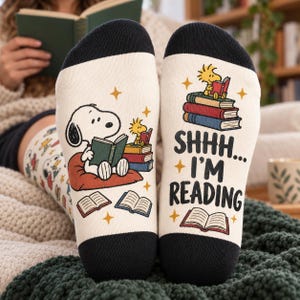 Op de afbeelding: Off-white sokken met zwarte teen- en hielstukken. De linkersok toont Snoopy lezend op een kussen, terwijl de rechtersok een stapel boeken met Woodstock erbovenop en de tekst SHHH... I'M READING bevat, versierd met kleine sterretjes.