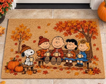 May include: A rectangular coir doormat featuring a printed autumn scene with Snoopy and the Peanuts gang sitting on a bench and a stool. They are holding steaming mugs under a large tree with orange and red leaves, surrounded by pumpkins.