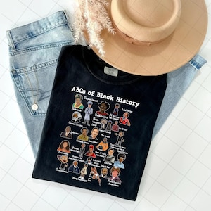 May include: Black t-shirt with the text "ABCs of Black History" and illustrations of historical figures. The shirt is paired with light blue jeans and a tan hat. A gold necklace and dried flowers are also visible.