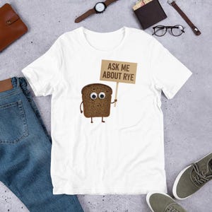 Könnte beinhalten: Weißes T-Shirt mit einem Cartoon-Roggenbrot-Charakter, der ein Schild mit der Aufschrift "ASK ME ABOUT RYE" hält. Das Brot hat Kulleraugen. Das Design ist auf dem T-Shirt zentriert.