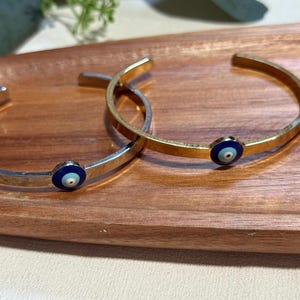 Könnte beinhalten: Zwei offene Armreifen, einer silberfarben und einer goldfarben, jeweils mit einem blauen und weißen Evil-Eye-Charm. Die Armreifen werden auf einem Holztablett präsentiert und zeigen ein minimalistisches Schmuckdesign.