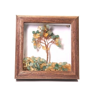 Puede incluir: Una escultura decorativa de árbol enmarcada. El árbol presenta un tronco y ramas de alambre de cobre adornados con piedras preciosas verdes y amarillas. La base del árbol está rodeada por un lecho de piedras preciosas similares, todo dentro de un marco de madera.