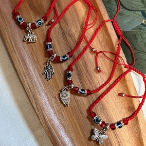 May include: Red string bracelets with adjustable closures. Each bracelet features a combination of blue evil eye beads, red beads, and a silver charm. Charms include an elephant, Hamsa hand, owl, and butterfly.