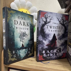 May include: Two hardback books, "One Dark Window" and "Two Twisted Crowns" by Rachel Gillig, are displayed on a wooden shelf. The first book features a dark castle illustration, while the second showcases a woman in a red cloak. Both books have the author's name prominently displayed.