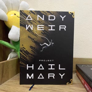 Puede incluir: Un libro de tapa dura titulado "Project Hail Mary" de Andy Weir. La portada presenta un fondo negro con pinceladas doradas y una ilustración de un astronauta cayendo. El título y el nombre del autor son en blanco.