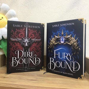 Op de afbeelding: Twee hardcover boeken, "Dire Bound" en "Fury Bound" van Sable Sorensen, worden getoond. Het eerste boek heeft een rood-zwarte omslag met een wolf en zwaard ontwerp. Het tweede boek heeft een blauwe omslag met een kroon en wolf ontwerp. Beide boeken hebben gouden hoekaccenten.