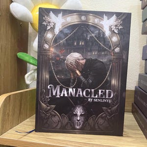 Op de afbeelding: Een hardcover boek getiteld "Manacled" door Senlinyu. De omslag toont een grijswaarden illustratie van een persoon met zijn gezicht in zijn handen, een gebouw en een masker. Het boek heeft een blauw bladwijzer.