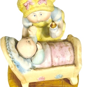 May include: A ceramic figurine depicting a doll-like figure feeding a baby in a crib. The figure has blonde hair with pink bows and is wearing a yellow dress. The baby is in a crib with a pink blanket. The base is a light brown color.