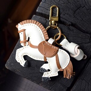 Puede incluir: Un llavero de caballo hecho de cuero blanco y marrón con un mosquetón y una anilla de metal dorado. La figura del caballo tiene una silla, brida y crines marrones, con cascos negros texturizados y una cola hecha de flecos de cuero.