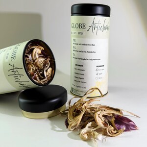 May include: Two cylindrical containers of Globe Artichoke tea, one open and spilling dried artichoke buds. The other container is upright with the label visible. The label includes text about the tea's benefits and preparation. A small pile of dried artichoke buds is in front.