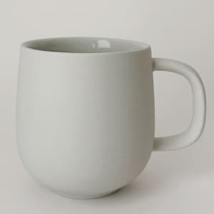 May include: A white ceramic mug with a rounded body and a curved handle. The mug has a matte finish and a smooth, clean design. The interior of the mug is a lighter shade of white.