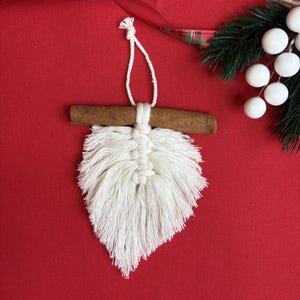 May include: A handmade macrame Christmas ornament with a cinnamon stick accent. The white, leaf-shaped ornament is suspended by a white cord. A red background with a plaid ribbon and white berries complete the festive scene.