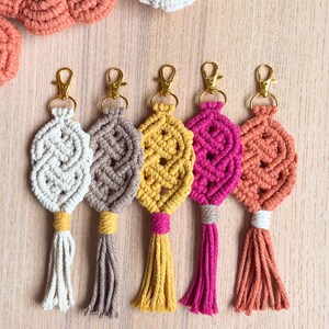 May include: Five macrame keychains in a row, each with a different color: off-white, brown, yellow, magenta, and orange. Each keychain has a gold-colored clasp and a matching tassel. The keychains are arranged on a light wood surface.