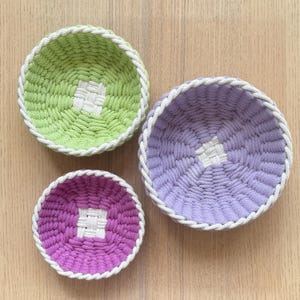 May include: Three handmade, round woven baskets in shades of green, purple, and pink. Each basket has a white woven center and a white rope trim. The baskets are arranged on a light wood surface.
