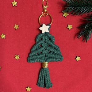 Może przedstawiać: Zielony brelok do kluczy w kształcie choinki z makramy, z drewnianą gwiazdką i złotymi akcentami. Brelok ma złoty pierścień i karabińczyk. Tło jest czerwone ze złotym konfetti w kształcie gwiazd.