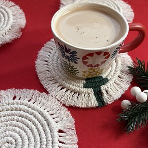 Könnte beinhalten: Eine Tasse heiße Schokolade mit rotem Henkel steht auf einem weißen Makramee-Untersetzer mit grünem und weißem Design. Weitere Makramee-Untersetzer und ein Zweig Grün mit weißen Beeren liegen auf einer roten Oberfläche.