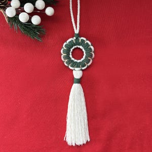 Può includere: Ornamento natalizio fatto a mano con un design a ghirlanda in macramè verde e bianco e un nappa bianca. L'ornamento è appeso a un filo bianco su uno sfondo rosso. Un rametto di verde con bacche bianche si trova nell'angolo in alto a sinistra.