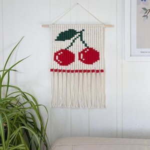 May include: A woven wall hanging featuring a pair of red cherries with green leaves against a white background. The design includes a red border and white fringe. The wall hanging is suspended from a wooden dowel.