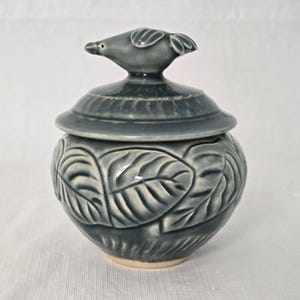 Könnte beinhalten: Graue Keramikdose mit Deckel und vogelartigem Griff. Die Dose ist mit Blattmustern verziert und hat einen hellen Sockel. Das Design ist ein charmantes, handgefertigtes Stück.