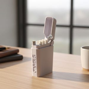 Puede incluir: Un estuche para cigarrillos de color gris claro con el nombre "JAMES" en relieve en el lateral. El estuche está abierto, revelando cigarrillos. Un bolígrafo y un cuaderno están a la izquierda, y una taza de café a la derecha.