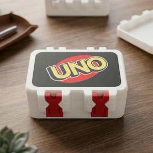 May include: White plastic UNO card game case with red clasps. The case has the UNO logo in yellow and red on a black background. Other cases and a tray are visible on a wooden surface.